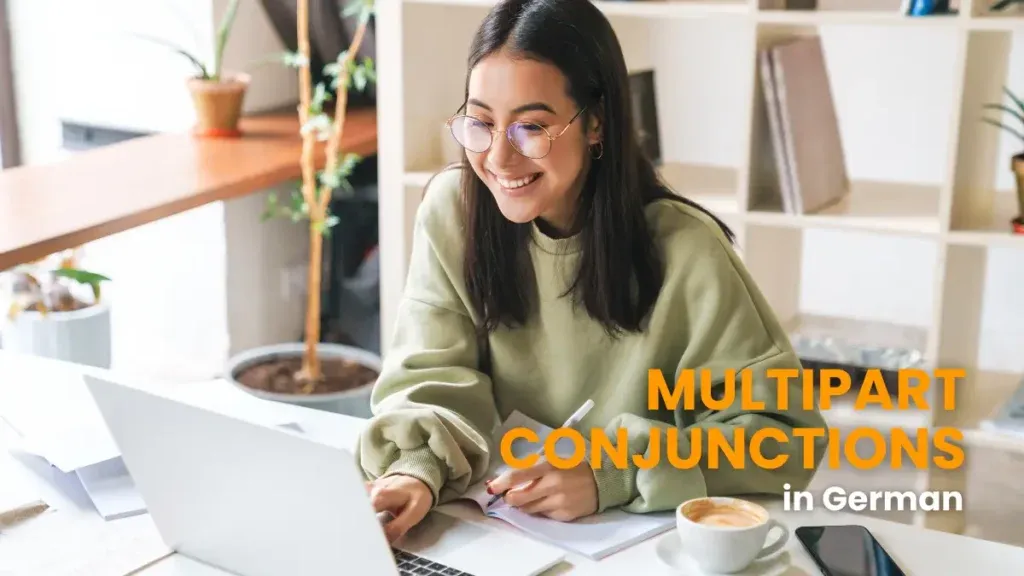 A smiling young woman wearing glasses and a green sweater sits at a desk working on a laptop, learning about multipart conjunctions in German.