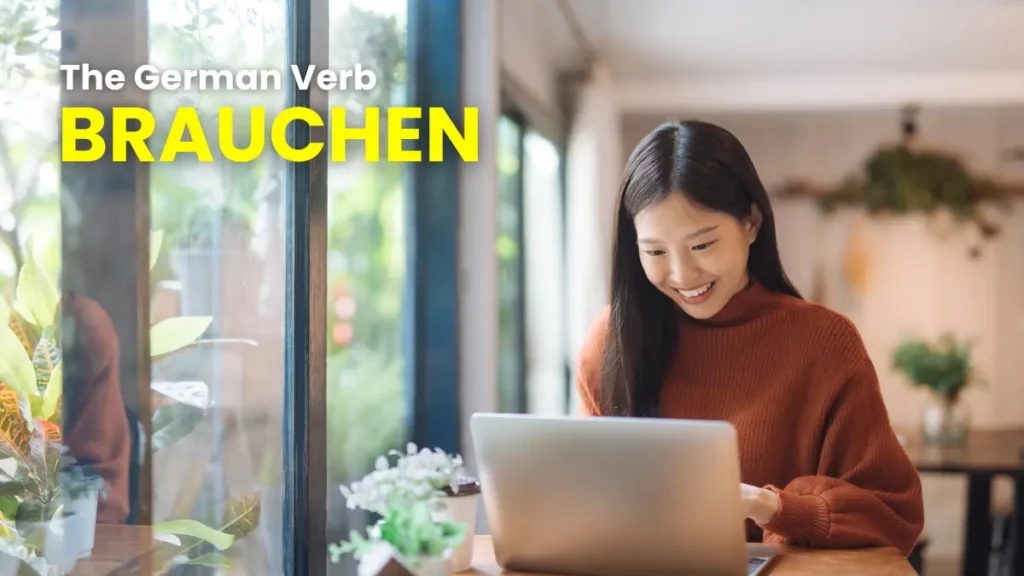 A young Asian woman wearing a red sweater smiles while using a laptop, learning about the German verb "brauchen".